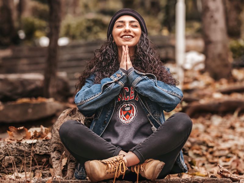 A woman sits cross-legged on the ground among trees, closing her eyes and smiling.