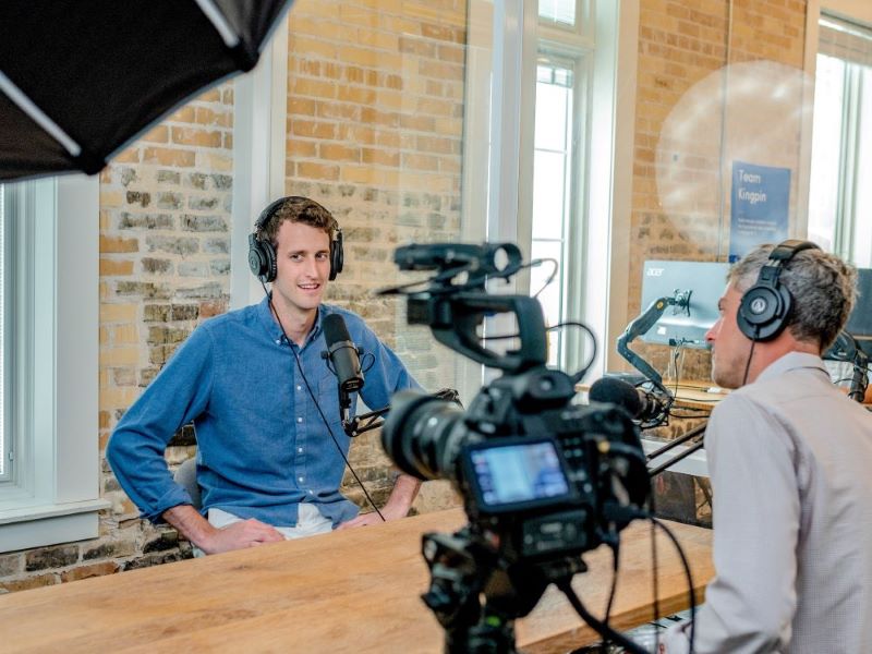 Two men sit at a table, speaking into microphones for a recorded interview.