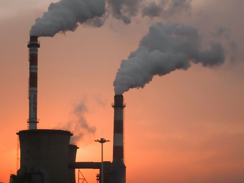 Smoke billows from a pair of factory chimneys, framed by a glowing orange sky.